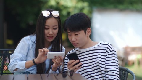 Tracking shot of teenage Asian girl using smartphone and discussing something on screen with friend while sitting at cafe table outdoors