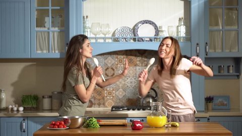 Portrait of two beautiful women dancing and singing at home kitchen using spoons, while cooking healthy breakfast with fresh vegetables and fruits slow motion