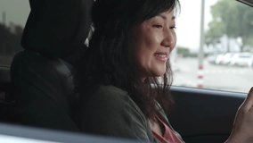 Tracking of mid-aged Asian woman smiling, waving at camera and speaking via video call on smart phone in car