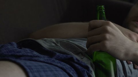 Man sleeping on couch with beer bottle, yawning and scratching belly, addiction