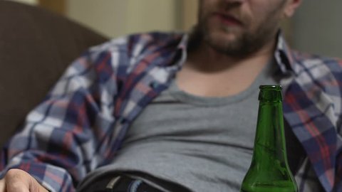 Drunk bearded man sitting on couch and drinking beer, addiction, bottom view