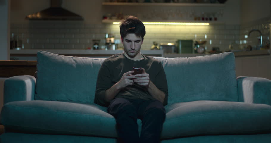 young man sitting on a sofa using his mobile phone not paying attention to the tv. Royalty-Free Stock Footage #1009173446