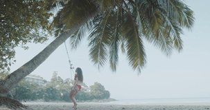 Beautiful young girl in swimsuit relaxing on the swing at the tropical beach - video in slow motion