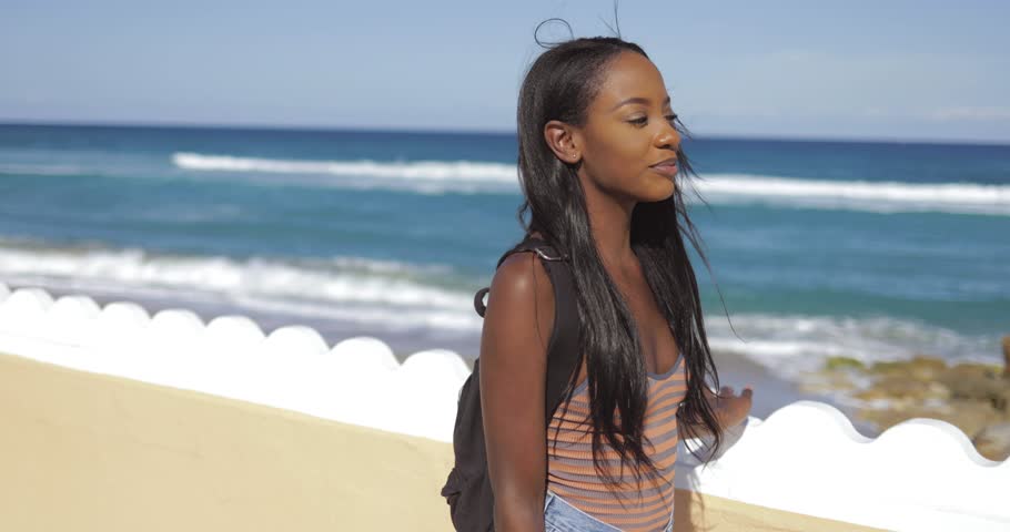 Back view of young ethnic pretty woman with backpack walking on pavement at the fence at seaside.