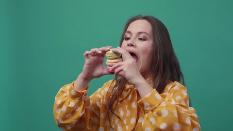 Beautiful hilarious woman wants to eat three macarons at the same time, widely opens her mouth, it doesn’t work out, tries to eat two of them, gives it up, looks upset. Dessert, delicious. Female