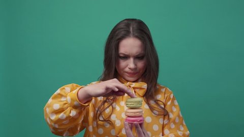 Pretty young woman puts three macarons one on top of another, wants to eat it all at the same time on a colorful background. Having fun, delicious dessert. Slow motion, female portrait