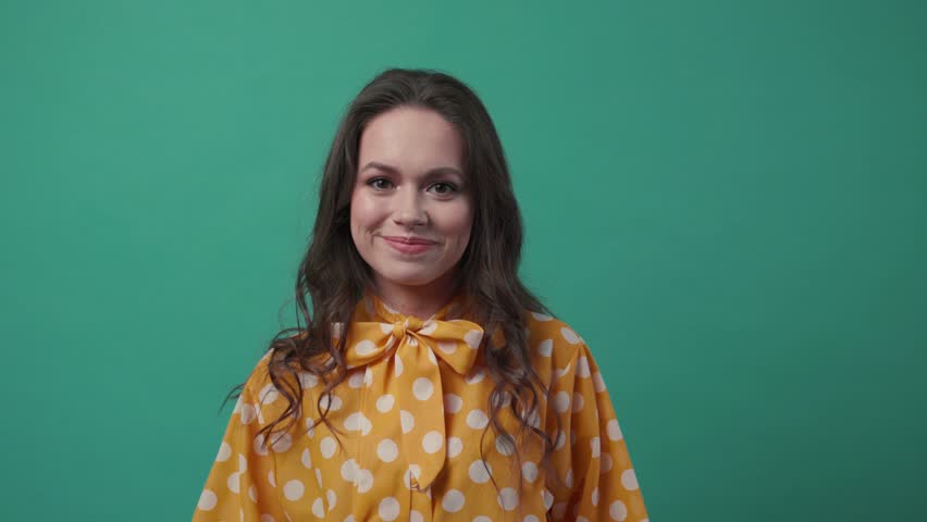 Beautiful young woman in a stylish blouse looks straight to camera and smiles happily on a green background. Happy moments, cheerful mood, positive emotions, being optimistic. Slow motion, female