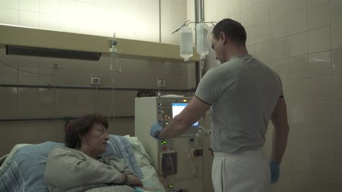 Male medical technician prepares hemodialysis machine for female patient treatment, pan left on the room with nurse and group of mature women that lying in the beds during peritoneal dialysis, indoors