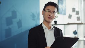 Happy asian young man with glasses hold documents look at camera smile show your finger up the hill in office chinese attractive handsome fashion men business leader businessperson company slow motion