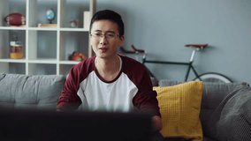 Happy smiling attractive young chinese man with glasses sits on the couch and turns on the TV at home looking house technology male business bored leisure video sofa portrait close up slow motion