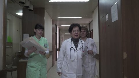 Two mature female doctors in white gowns with stethoscope around neck and younger nurse hold documentation walk through the hall of hospital building and enter in room with patients, steady cam shot