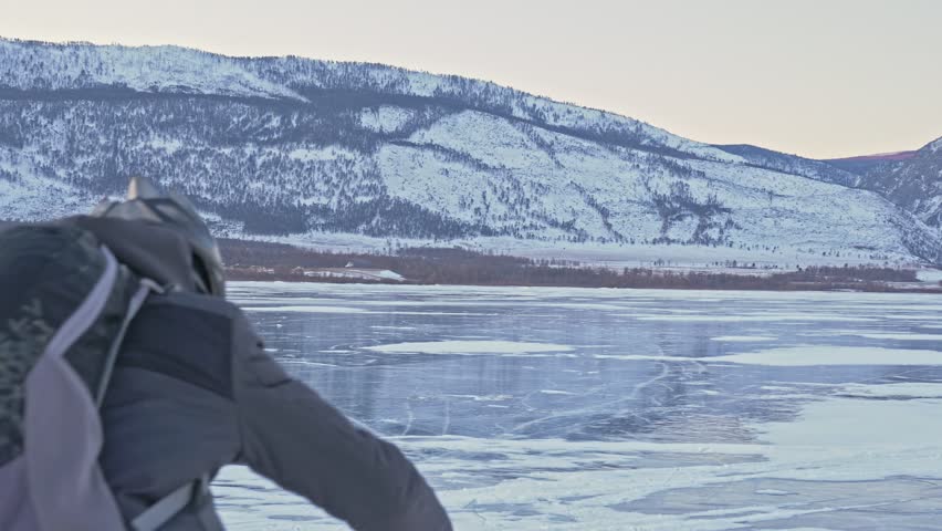 Man is riding a bicycle on ice. The cyclist is dressed in a gray down jacket, backpack and helmet. Ice of the frozen Lake Baikal. The tires on the bicycle are covered with special spikes. The traveler