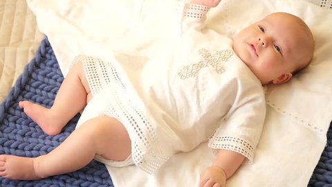 A cute kid lies on a bed in a shirt for baptism, moves his arms and legs, studies the world around, smiles, looks at the camera. HD, 1920x1080. slow motion.