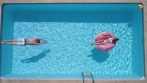 Aerial - Adult man dives into the the pool while girl is lying on a donut pool float (slow motion)