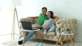 Joyful millennial couple streaming video content on line using laptop pc while sitting comfortable on the sofa in domestic room. Smiling positive couple watching tv show online on notebook pc at home.