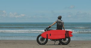 Handsome man biker surfer standing next to black motorbike cafe racer with red surfboard shortboard on beach at sunny day and looking waves. 4k video shooting by handheld gimbal