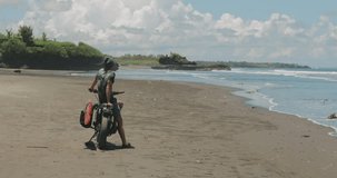 Handsome man biker surfer standing next to black motorcycle cafe racer with red surfboard shortboard on beach at sunny day and looking waves. 4k video shooting by handheld gimbal