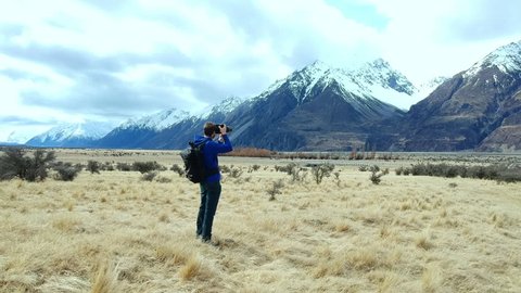 Young adventure photographer alone taking photographs around Mt Cook region of New Zealand. Drone shot tracks forward past man with snow capped mountains in background.