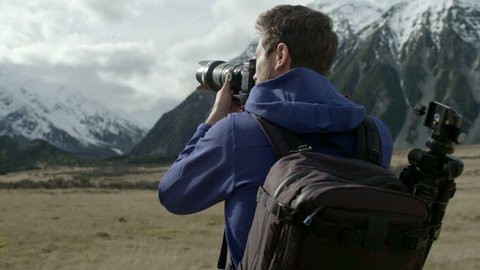 Young adventure photographer taking photographs around Mt Cook, New Zealand. Camera moves around man in slow motion as sun peeks through clouds.