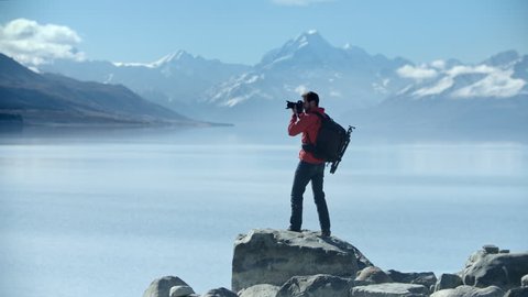 Young adventure photographer climbs atop a rock to take photos of the spectacular view with Mt Cook in distance. Mountain view of Lake Pukaki, New Zealand. Shot in 10bit Pro Res