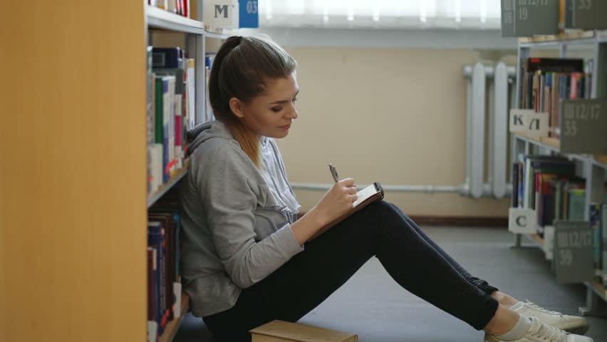 Young beautiful dreamy student girl is sitting on floor in lighty library among bookshelves writing down composition in copybook looking up at ceiling