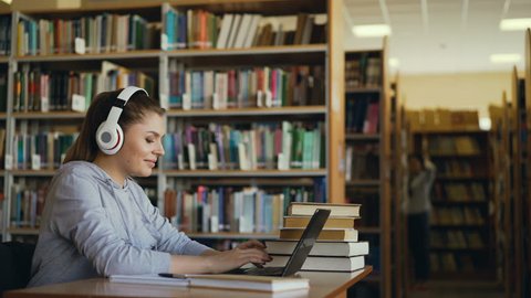 Beautiful positive caucasian female student with big headphones working at table in spacious library in front of laptop. She is smiling and texting