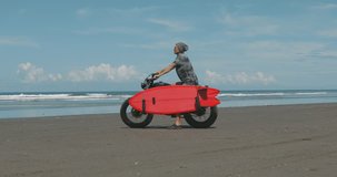 Handsome man biker surfer sitting on black motorbike cafe racer with red surfboard shortboard on the beach at sunny day and looking at waves. 4k video shooting by handheld gimbal