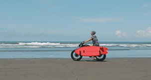 Handsome man biker surfer starts moving on black motorbike cafe racer with red surfboard shortboard on the beach along the ocean at sunny day. 4k video shooting by handheld gimbal