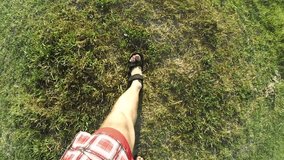 Feet in sandals with legs in shorts walking on earth and green grass in summer - view from above.