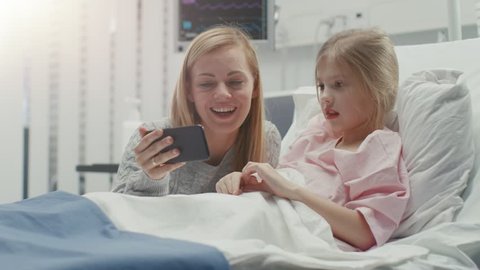 Cute Little Girl Lies on a Bed in the Children's Hospital, Her Mother Sits Beside, They Watch Cartoons/ Funny Videos on Smartphone. Modern Pediatric Ward.