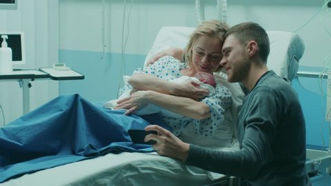 Father Takes Selfie of Him with His Wife Holding Newborn Babie while Lying on the Hospital Bed. Happy Young and Smiling Family. Shot on RED EPIC-W 8K Helium Cinema Camera.
