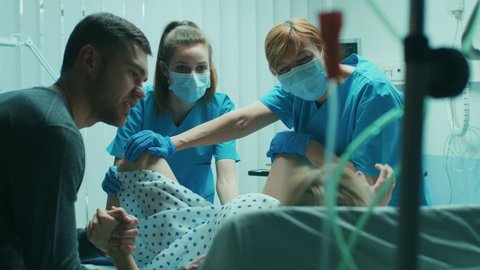 In the Hospital Woman in Labor Pushes to Give Birth, Obstetricians Assisting, Husband Holds Her Hand for Support. Modern Maternity Ward with Professional Midwives. Shot on RED EPIC-W 8K Helium Camera.