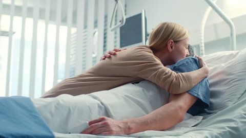 In the Hospital, Happy Wife Visits Her Recovering Husband who is Lying on the Bed. They Lovingly Embrace and Smile. Shot on RED EPIC-W 8K Helium Cinema Camera.