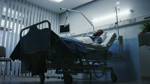 Low Level Shot in the Hospital, Very Sick Man Lying on the Bed, Nurse Checks His Vital Signs and Drop Counter. Shot on RED EPIC-W 8K Helium Cinema Camera.