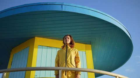Cute young woman in yellow raincoat standing on lifeguard station at sea at miami beach.