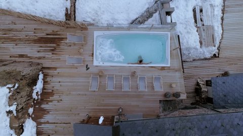 Aerial drone shot of man relax and chill on cold winter day in steaming hot water of tub or swimming pool, in luxurious expensive hotel or spa with wood floor and snow around