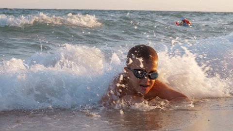 Slow motion shot of a boy in goggles lying on the shore and enjoying sea waves rolling in and washing him, scene at sunset. Summer vacation at the seaside