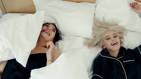 Close up shot of a two attractive lesbian girls falling onto bed with pillows. They are laughing hard and looking at each other.