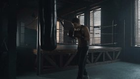 Lightweight boxer training in the dark vintage stylized gym. Caucasian young man with boxing gloves hitting the punching bag hard near the old boxing ring. Slow motion video.
