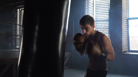Lightweight boxer training in dark vintage stylized gym. Caucasian young man with boxing gloves hitting the punching bag near the old boxing ring. 4k video.