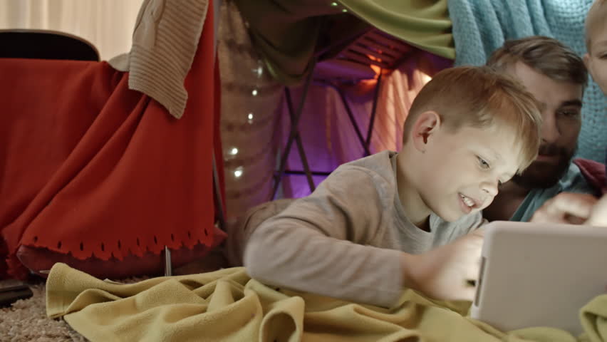 Tracking shot of happy and two little sons lying on carpet under play tent and playing together on digital tablet
