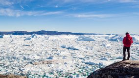 Travel in arctic landscape nature with icebergs - Greenland tourist man explorer - tourist person looking at amazing view of Greenland icefjord - aerial video. Man by ice and iceberg in Ilulissat.
