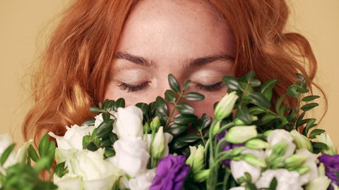 Portrait of redhead feminine woman holding colorful bouquet and enjoying smell of lisianthus flowers with closed eyes, isolated over beige background Adlı Stok Video