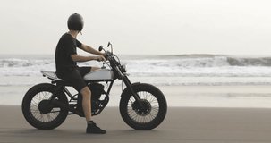 Handsome man biker driving his motorcycle caferacer on beach along ocean during sunset. 4k video shooting by handheld gimbal