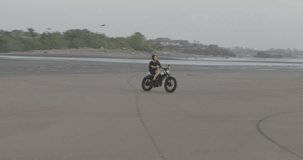 Handsome man biker driving his motorcycle cafe racer on the beach along the ocean during sunset. 4k video shooting by handheld gimbal