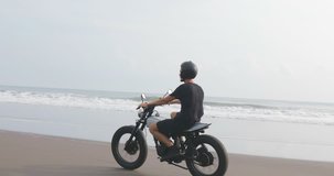 Handsome man biker driving his black motorcycle cafe racer on the beach along the ocean during sunset. 4k video shooting by handheld gimbal