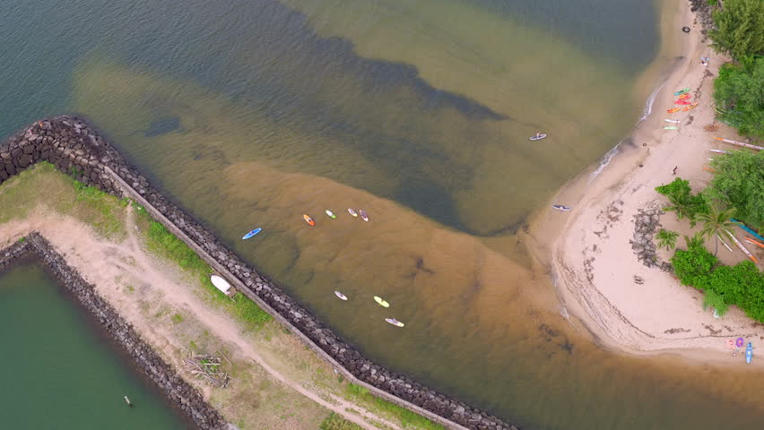 Slow push-in shot from a drone of people paddle boarding along a break wall in the mouth of a river in Hawaii, featuring colorful palms off to the side and textured sandbars. 