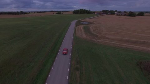 Aerial view of red car driving on country road. Cinematic drone shot flying over road and fields in Southern Sweden