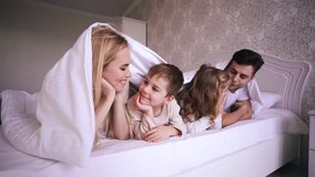 Young pretty happy parents and children talking with each other and lying under blanket in bedroom