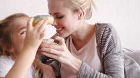 Young blonde pregnant mother eating with her funny pretty daughter donuts while sitting on sofa 